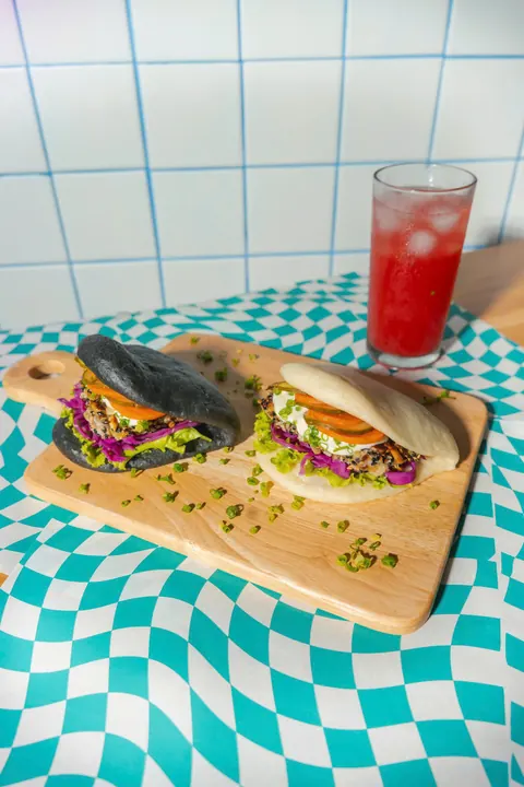 A black bao and a white bao on a wooden board on a turquoise checked cloth, with a red iced drink