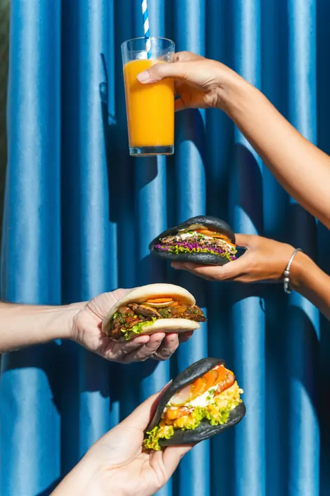 Hands holding three bao and a glass of orange juice against a blue curtain