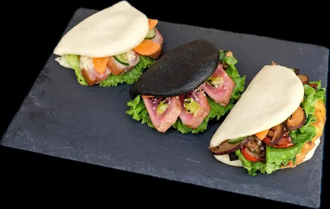 Three gua bao on a slate board, the middle one a black bun filled with slices of seared pink tuna