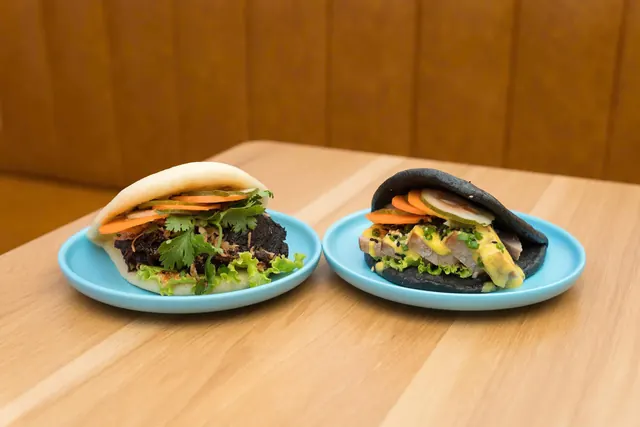 Two gua bao on blue plates at Bao Bao, one in a white steamed bun and one in a black bun